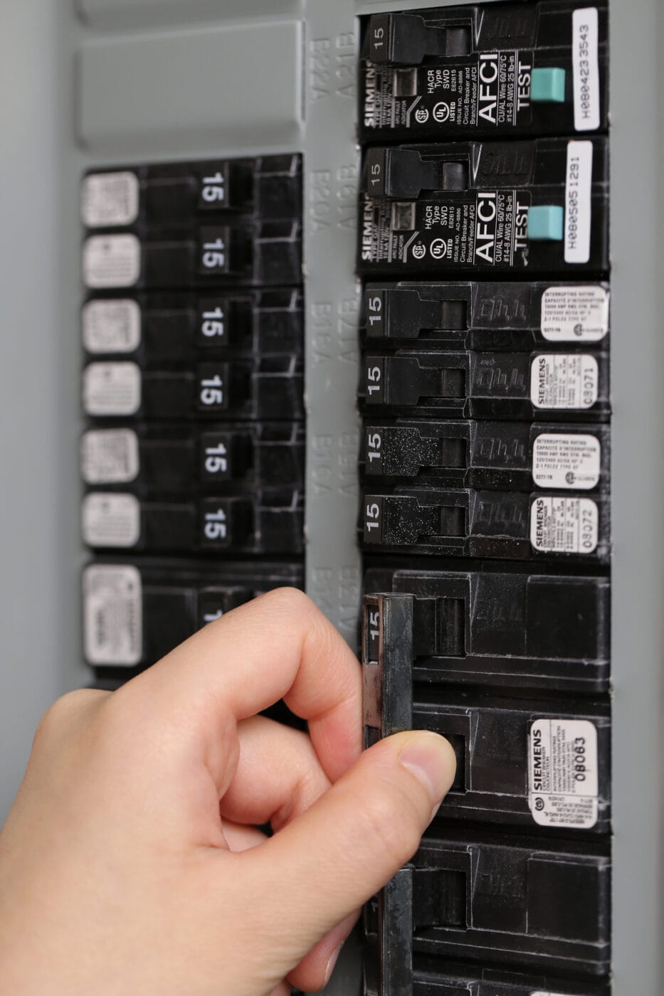 Hand adjusting a switch on a circuit breaker panel labeled with multiple circuit breakers, each marked with numbers and labels.