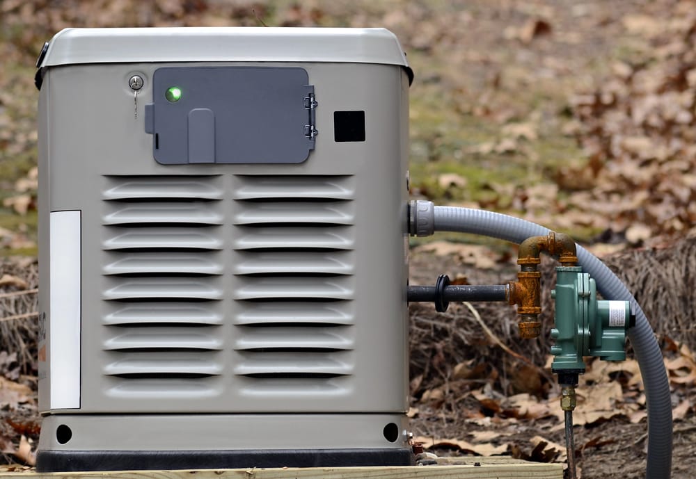 A standby generator with a green indicator light is connected to a gas line outdoors, surrounded by dry leaves and grass.