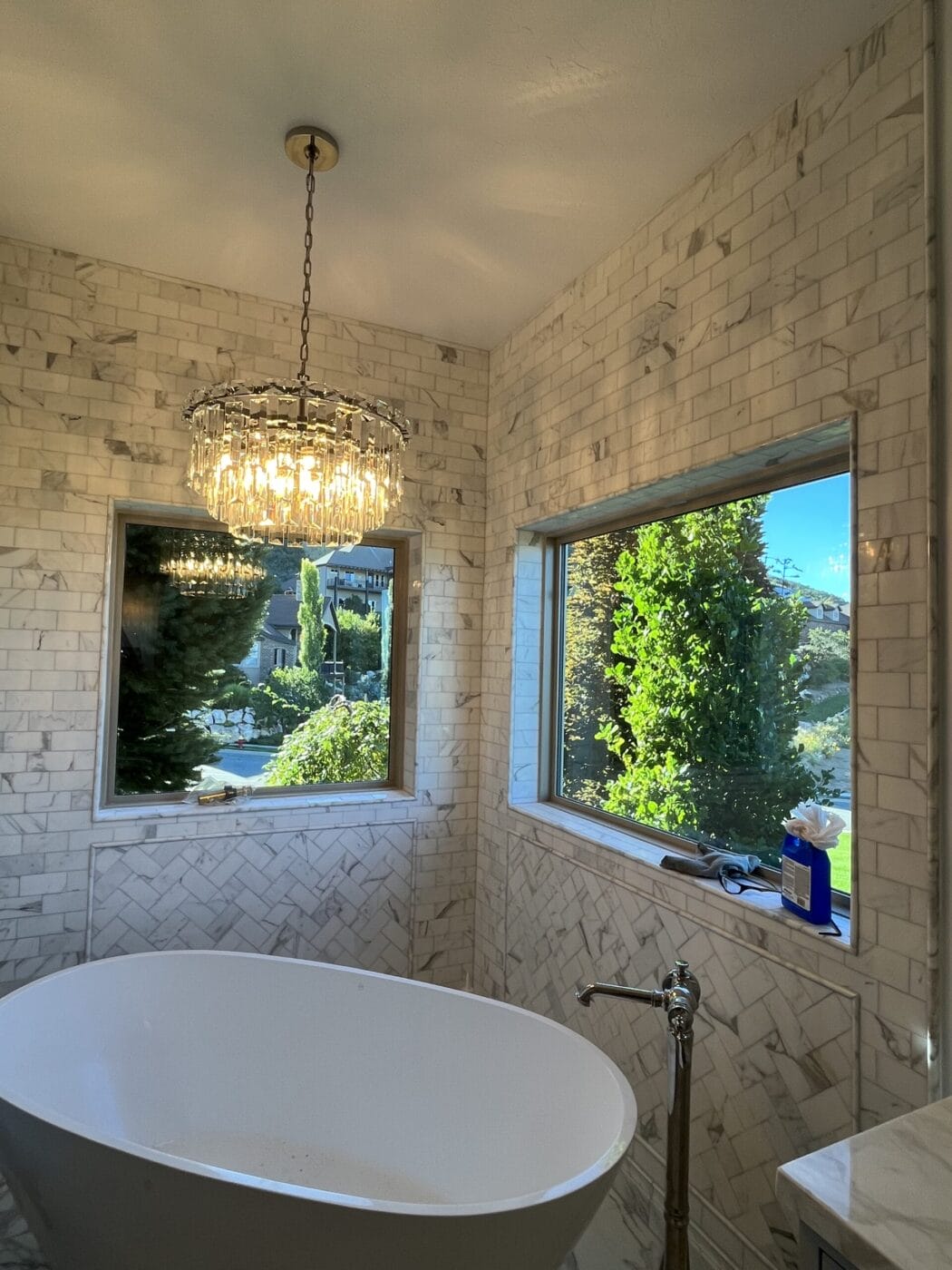 A bathroom with white tiled walls, a freestanding oval bathtub, a window view of greenery, and a chandelier above.