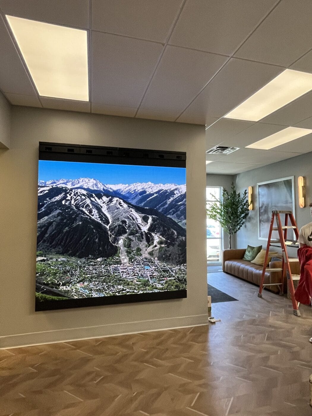 Large wall-mounted screen displaying an aerial view of a mountainous landscape with a ski resort. Indoors, a ladder and a couch are visible nearby.
