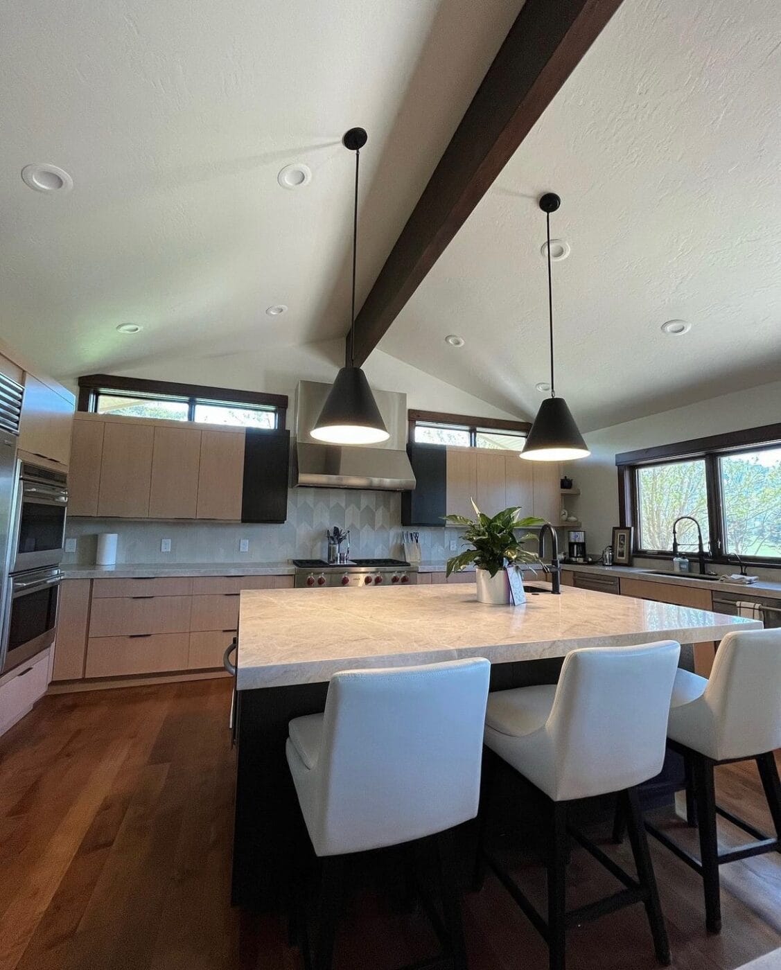 Modern kitchen with wooden floors, an island with white chairs, black pendant lights, and large windows. Light wood cabinets and stainless steel appliances are visible.