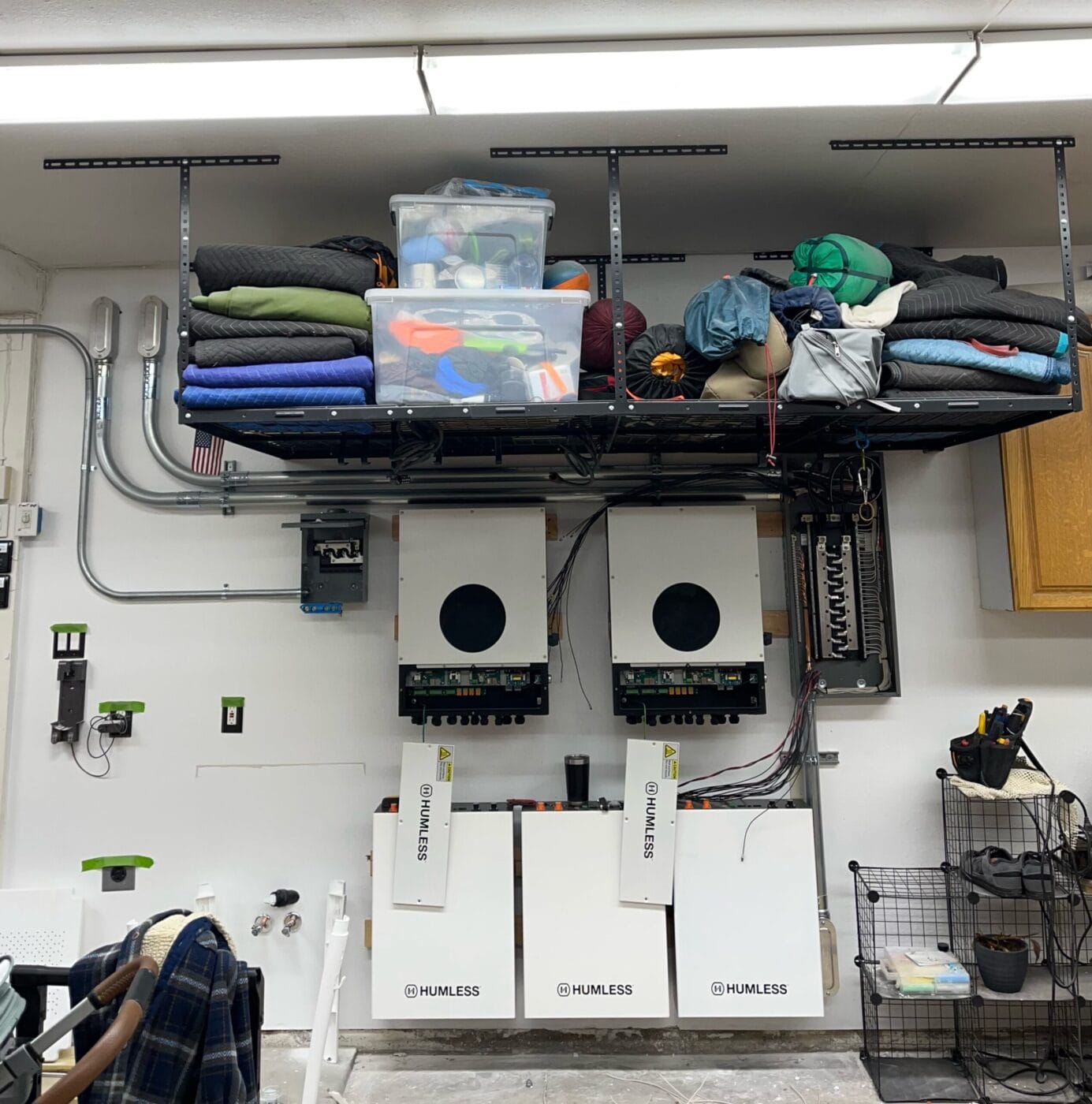 Garage with three wall-mounted power inverters, storage boxes, and camping gear on a shelf above. Cables and conduits are visible, along with a wire basket and a wooden cabinet on the side.