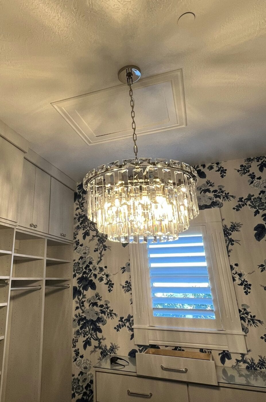 A stunning crystal chandelier serves as the centerpiece of this elegant room, showcasing sophisticated lighting installation. The space features floral wallpaper, white cabinetry, and a window adorned with classic white shutters.