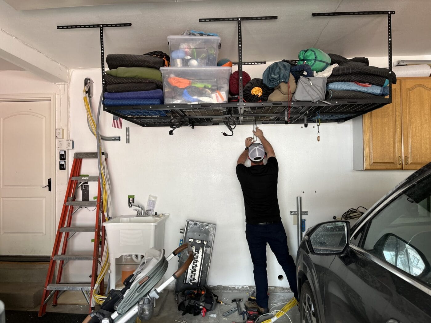 Man organizing items on a high garage shelf. Various tools and boxes are visible. A ladder and a car are nearby.