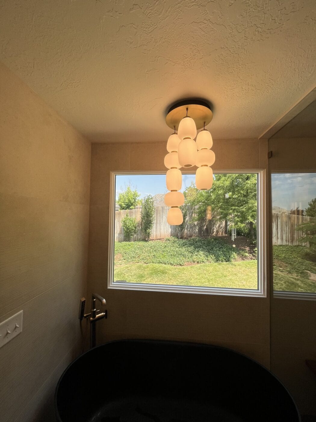 Bathroom with a black bathtub, modern pendant light, large window showing a fenced yard with trees and grass outside.